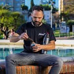 A man sits by a fountain preparing a traditional mate beverage in a sunny park setting.